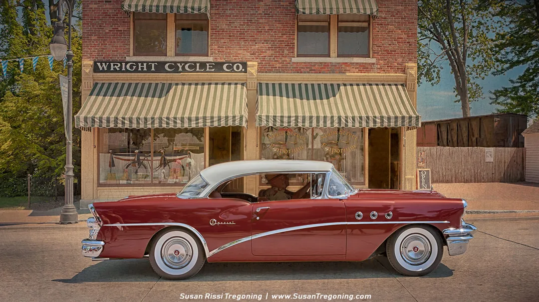 A red and white vintage Buick is parked in front of the historic Wright Cycle Co. brick storefront, which features green‑and‑white striped awnings and large display windows filled with bicycles. The early‑20th‑century setting creates a nostalgic scene tied to the Wright brothers’ bicycle shop. 