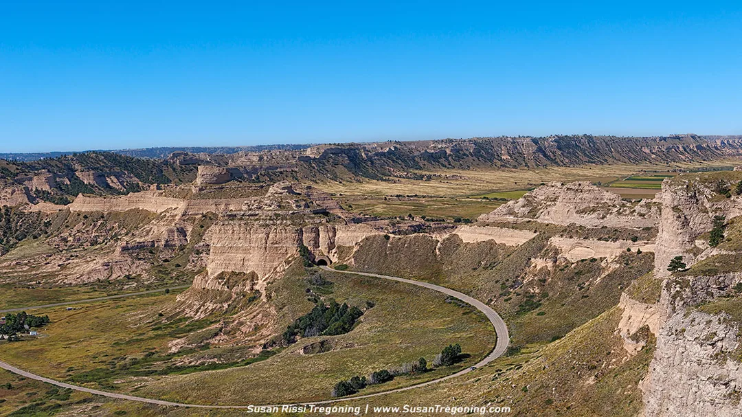   
A winding paved road curves through rugged cliffs and eroded rock formations on the ascent to the top of Scotts Bluff, with open plains and distant ridges visible under a clear blue sky.