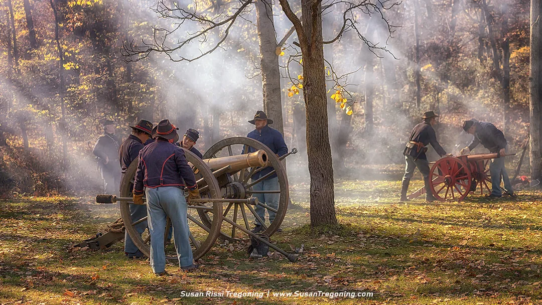 Civil War reenactors in Union uniforms operate two cannons in an autumn woodland setting. Sunlight filters through the trees as smoke drifts across the scene, with one group gathered around a large cannon in the foreground and another working with a smaller red‑wheeled cannon in the background.