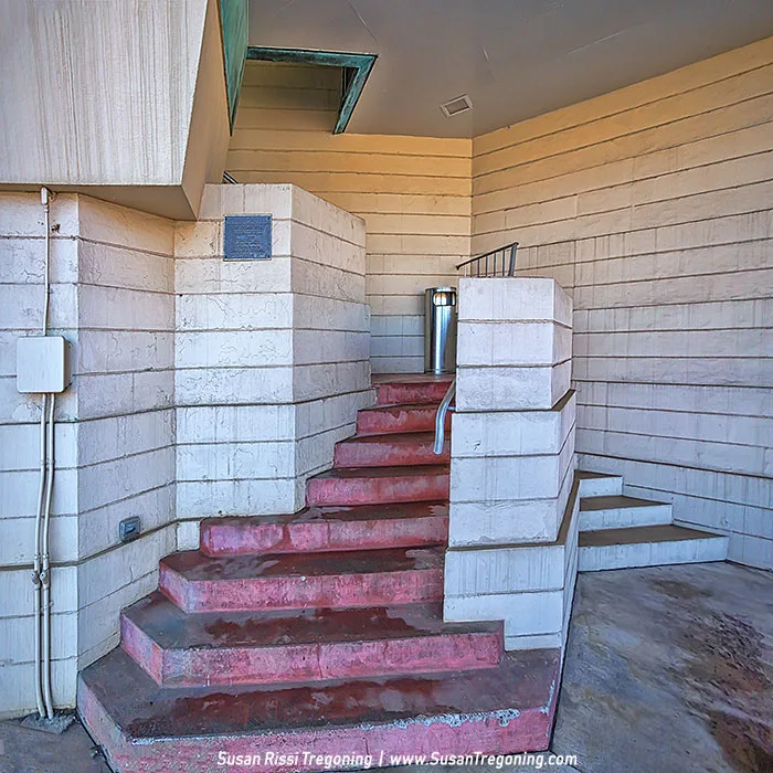 A stairway with red concrete steps splits in two directions, one set rising straight ahead and another curving to the right. Beige walls with horizontal lines frame the space, and a metal handrail runs along the central wall. A cylindrical metal trash can sits at the top of the stairs, with a small wall plaque and visible electrical conduits nearby. A recessed ceiling light and a green‑tinted window or vent complete the mid‑century architectural scene.