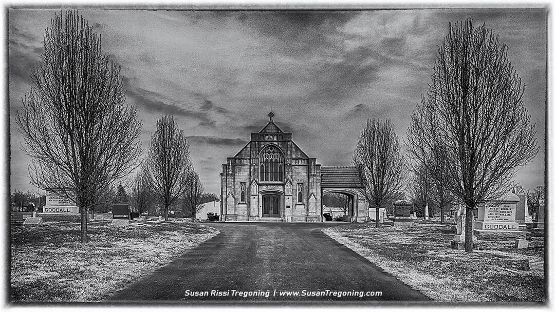 A black‑and‑white photograph of the historic Goddard Chapel in Marion, Illinois, centered at the end of a straight paved walkway in Rose Hill Cemetery. Leafless trees line both sides of the path, with rows of gravestones visible beneath them. Two matching headstones in the foreground read “GOODALL.” The Gothic‑style stone chapel rises in the distance with pointed arches and ornate window tracery, framed by a cloudy, dramatic sky.