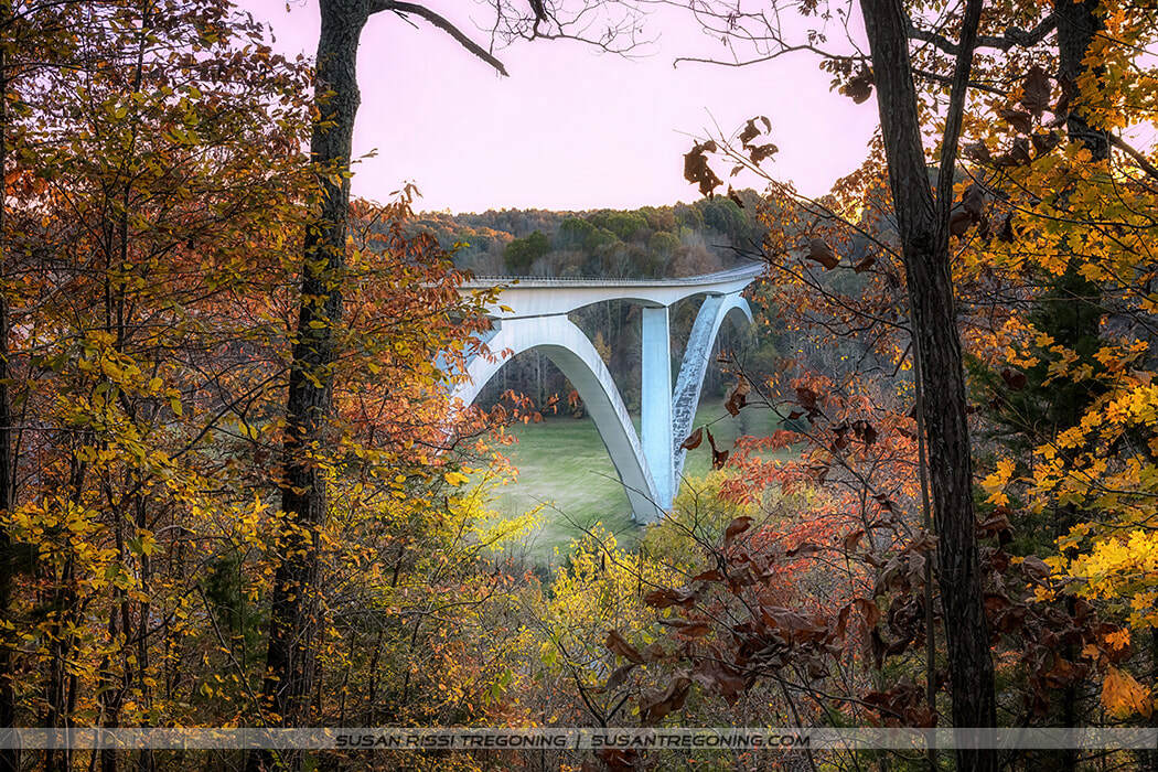 Double‑arched concrete bridge spanning Birdsong Hollow on the Natchez Trace Parkway, shown after sunset in autumn. The 1994 structure rises high above the valley with its two sweeping arches visible against the surrounding hills.