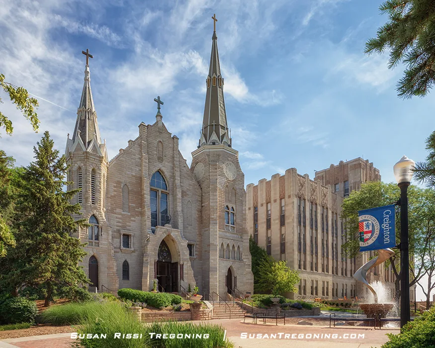A tall English Gothic church built of light stone stands at the center of the University Mall at Creighton University. The façade features a prominent square tower with pointed‑arch openings, narrow lancet windows, and strong vertical lines. Mature trees and campus walkways frame the building, and the sky above is clear and bright.