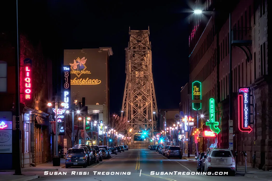   
A winter street scene in Duluth shows the Aerial Lift Bridge at night, with snow on the ground and very few cars or people out in the extreme cold.
