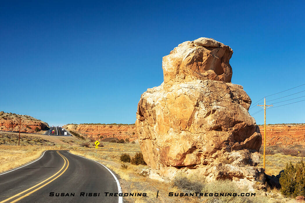 A paved road curves toward a tan rock formation on the right side. The rock has a rounded top and vertical face, with dry grasses and shrubs at its base. Additional cliffs and a bridge with vehicles are visible in the distance under a clear blue sky.