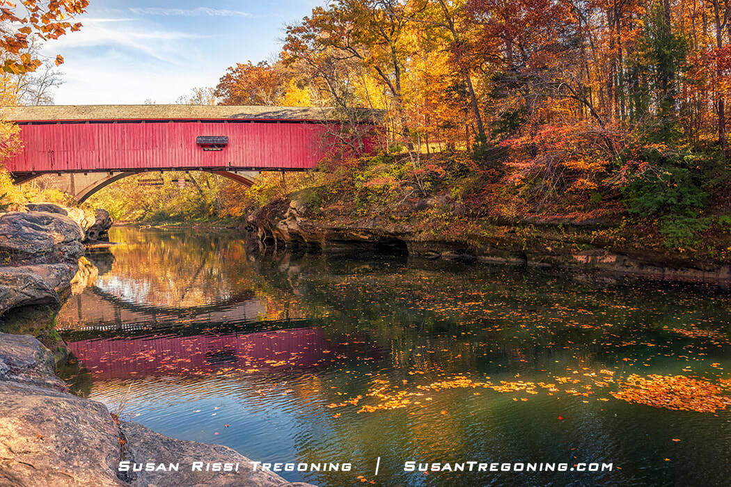 A red covered bridge spans Sugar Creek at Turkey Run State Park, reflected in the calm water surrounded by bright autumn foliage and large rocks along the bank.