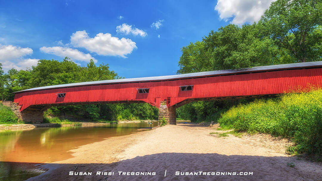 Sugar Creek flowing beneath the long red West Union Covered Bridge, with stone piers, sandy banks, and lush green trees under a bright blue sky.