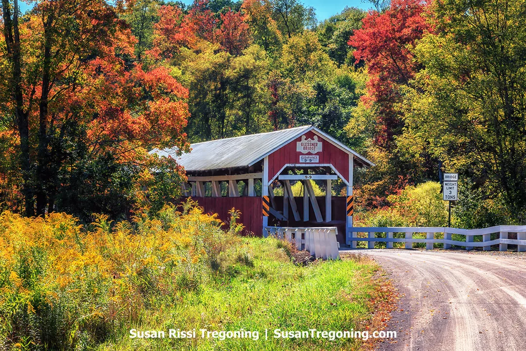 A red covered bridge with white trim stands along a narrow rural road surrounded by autumn trees in shades of red, orange, yellow, and green. A sign above the entrance reads Glessner Covered Bridge with a posted clearance of 9 feet 9 inches. A separate roadside sign to the right lists a bridge weight limit of 3 tons.