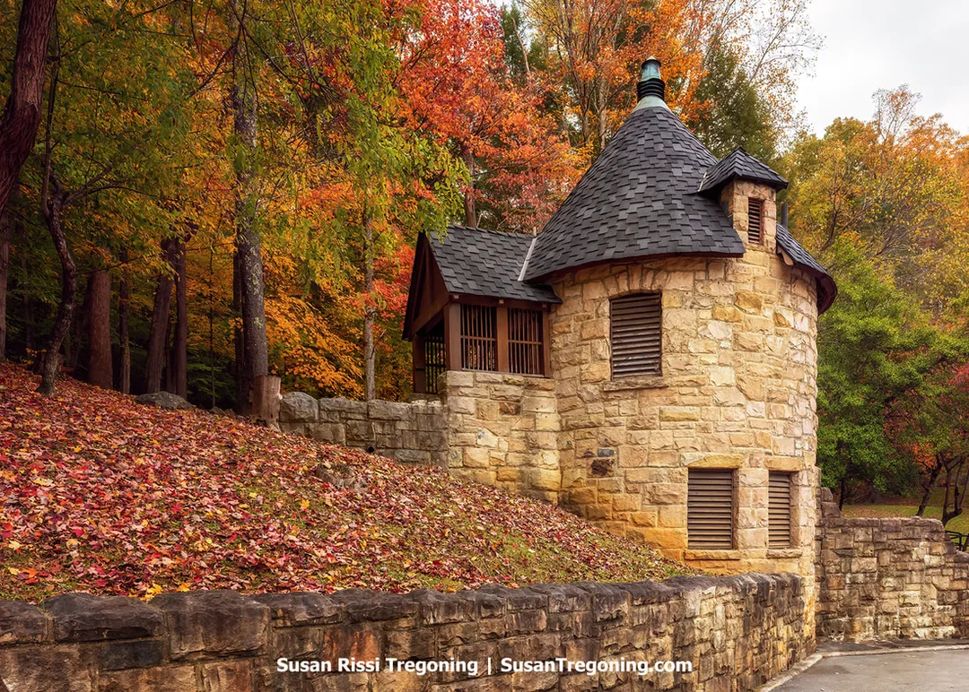 A small two‑tiered round stone tower with a conical roof stands beside a stone wall, surrounded by autumn trees with red, orange, and yellow foliage and fallen leaves on the ground.