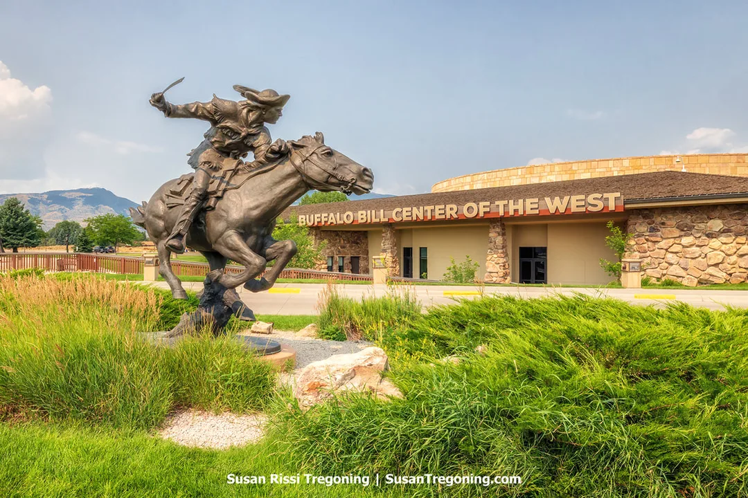 A bronze statue of a rider on a galloping horse positioned in front of a building labeled Buffalo Bill Center of the West, with landscaped greenery and mountains in the background.
