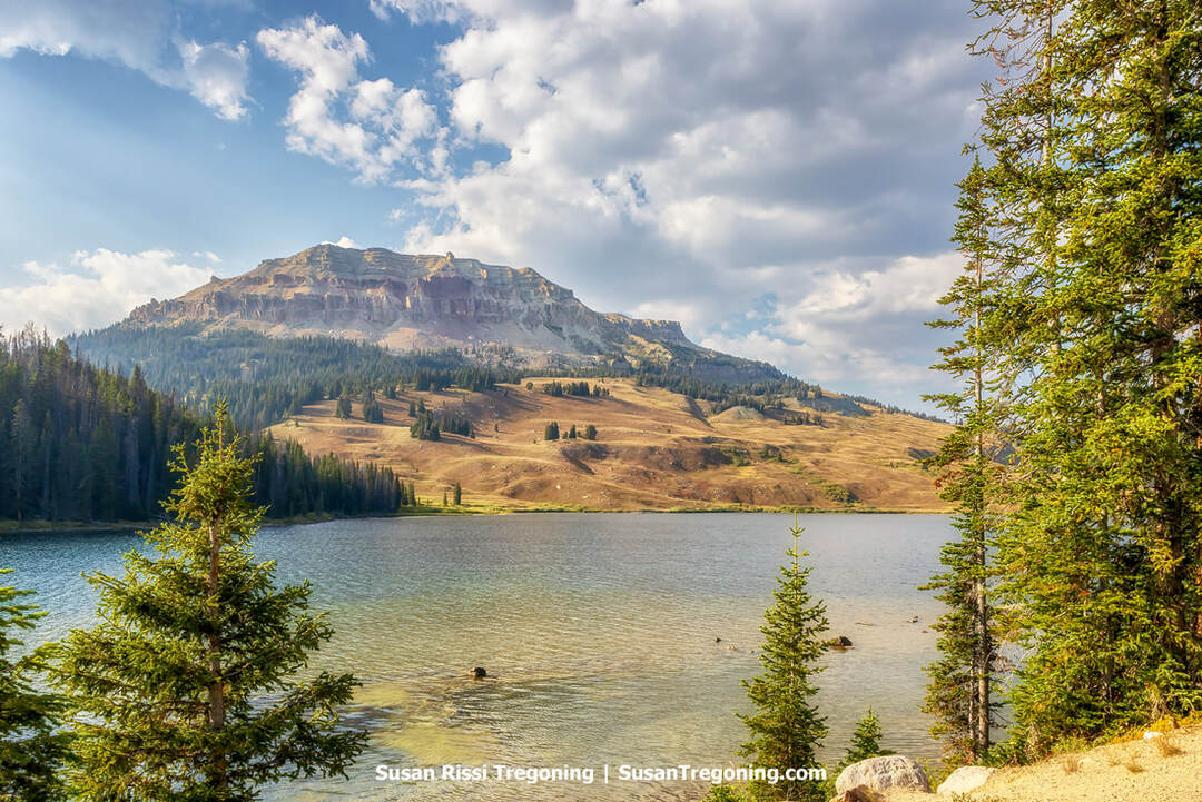A calm mountain lake bordered by evergreen trees, with a large rocky butte rising behind it. Layered rock formations, forested slopes, and open grassy areas appear beneath a partly cloudy sky.