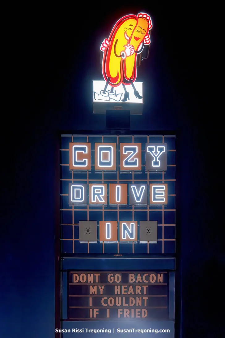 The new Cozy Drive‑In sign is lit at night on Route 66 in Springfield, Illinois. The sign features the hugging Cozy Dog characters at the top and retro 1970s Googie‑style lettering spelling Cozy Drive In. The message board below displays the phrase: Don't go bacon my heart I couldn't if I fried.