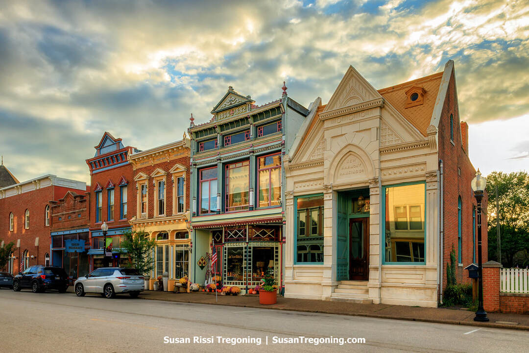 Historic storefronts along the 500 block of Main Street in New Harmony include the New Harmony Banking Company and the Mumford Emporium, both housed in ornate nineteenth‑century brick buildings. Decorative façades, arched windows, and detailed trim line the streetscape, with parked cars, potted plants, and American flags adding small touches of color beneath a partly cloudy sky.