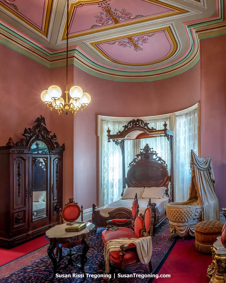A richly decorated Victorian bedroom features patterned wallpaper, an ornate wooden bed with a carved headboard, and a matching dresser with a large mirror. A chandelier hangs from the ceiling, and antique furnishings and décor fill the elegant historic room.