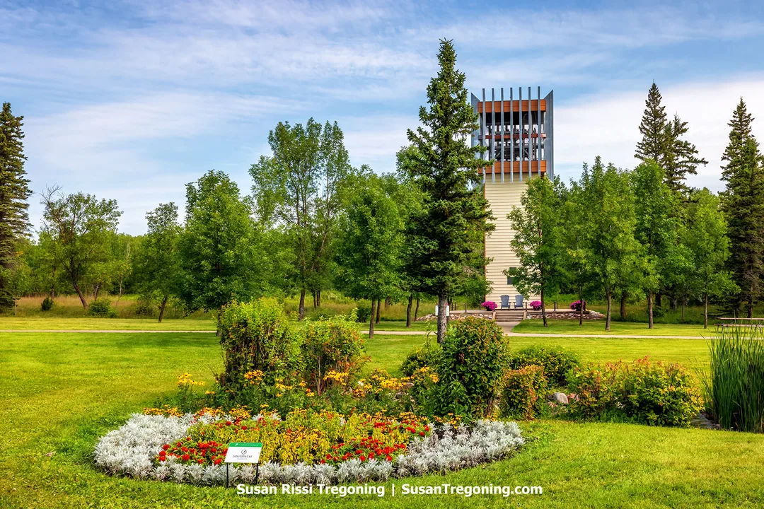 The Carillon Bell Tower at the International Peace Garden stands tall among green trees and manicured lawns along the North Dakota–Canadian border. A colorful flower bed blooms in the foreground, symbolizing unity and friendship between nations within this 2,300‑acre garden dedicated to peace and harmony.