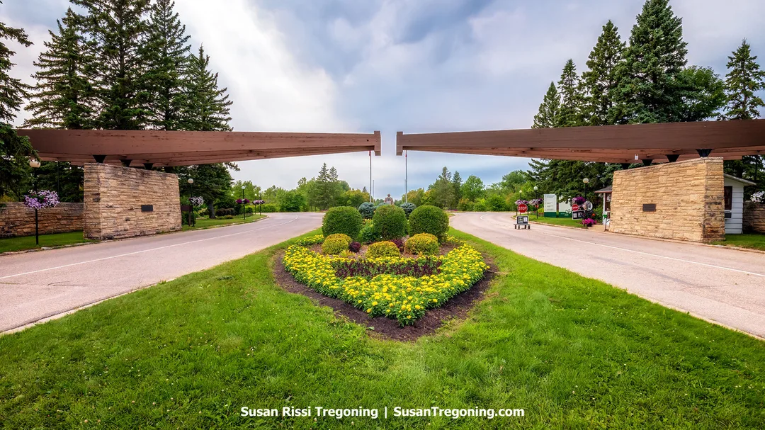 A stone‑and‑timber entrance structure at the International Peace Garden with the United States pylon on the left and the Canada pylon on the right, each supporting angled wooden beams that reach toward one another above a heart‑shaped flower bed.