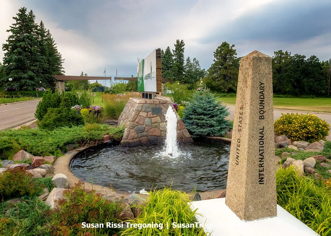 A granite International Boundary Marker installed in 1932 stands on the 49th parallel at the entrance of the International Peace Garden, marking the historic border established by the 1818 Treaty between the United States and Canada. A fountain symbolizes renewal beside it, and in the distance the Garden’s cantilevered entrance aligns its open center precisely with the boundary line, reinforcing the site’s theme of binational cooperation.