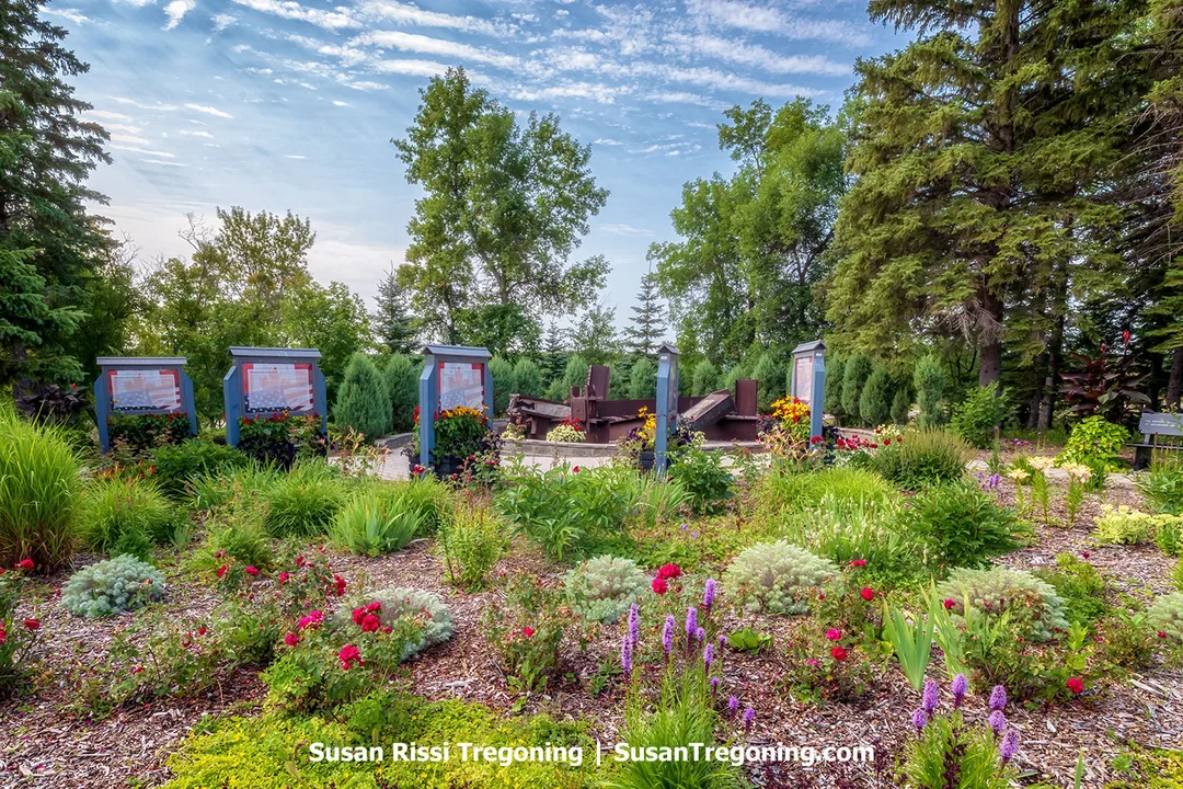 Steel girders recovered from the World Trade Center stand upright within the 9/11 Memorial at the International Peace Garden, surrounded by bright summer flowers and evergreens. The weathered beams rise as solemn reminders of the September 11 attacks, placed here in 2002 to honor the victims and symbolize the shared commitment to peace between the United States and Canada.