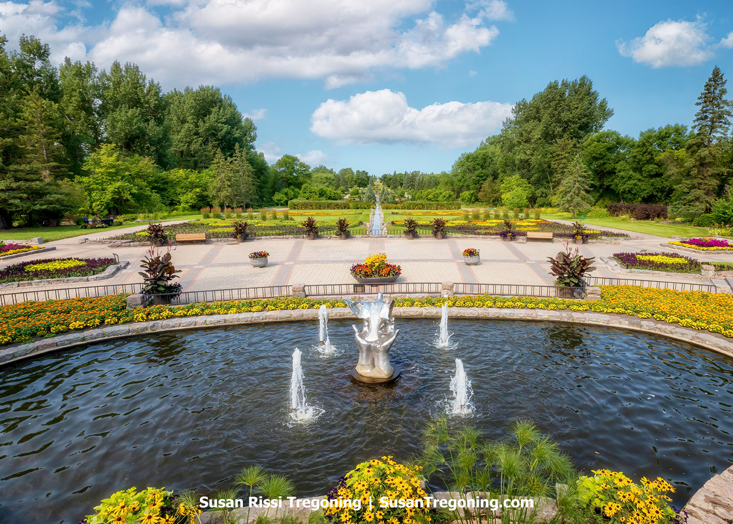 View of the International Peace Garden with a central fountain, colorful flowerbeds, tree-lined walkways, and the small border river marking the U.S.–Canada boundary under a bright summer sky.