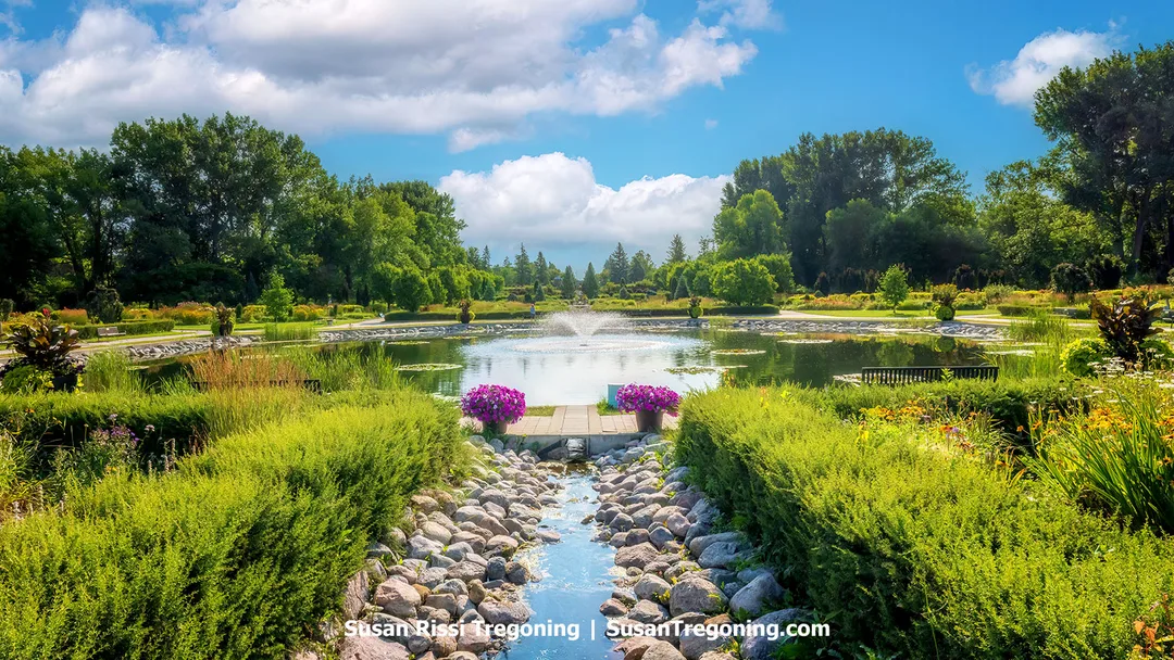 Sunken Garden Fountain Pool at the International Peace Garden with the stone‑lined international border channel in the foreground, surrounded by colorful flowers, manicured hedges, and a bright summer sky