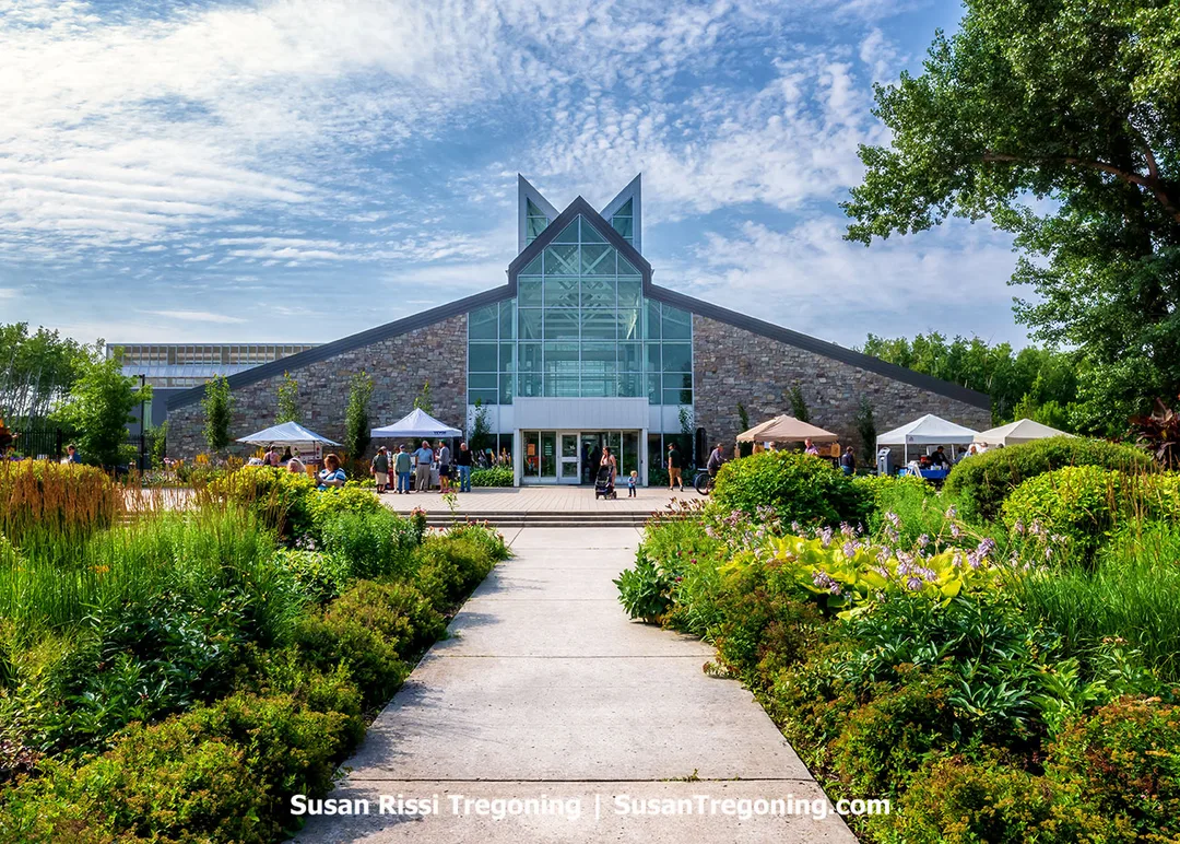 Conservatory and Interpretive Centre at the International Peace Garden, featuring a modern glass‑and‑stone design with white canopy tents and vibrant summer gardens in the foreground.