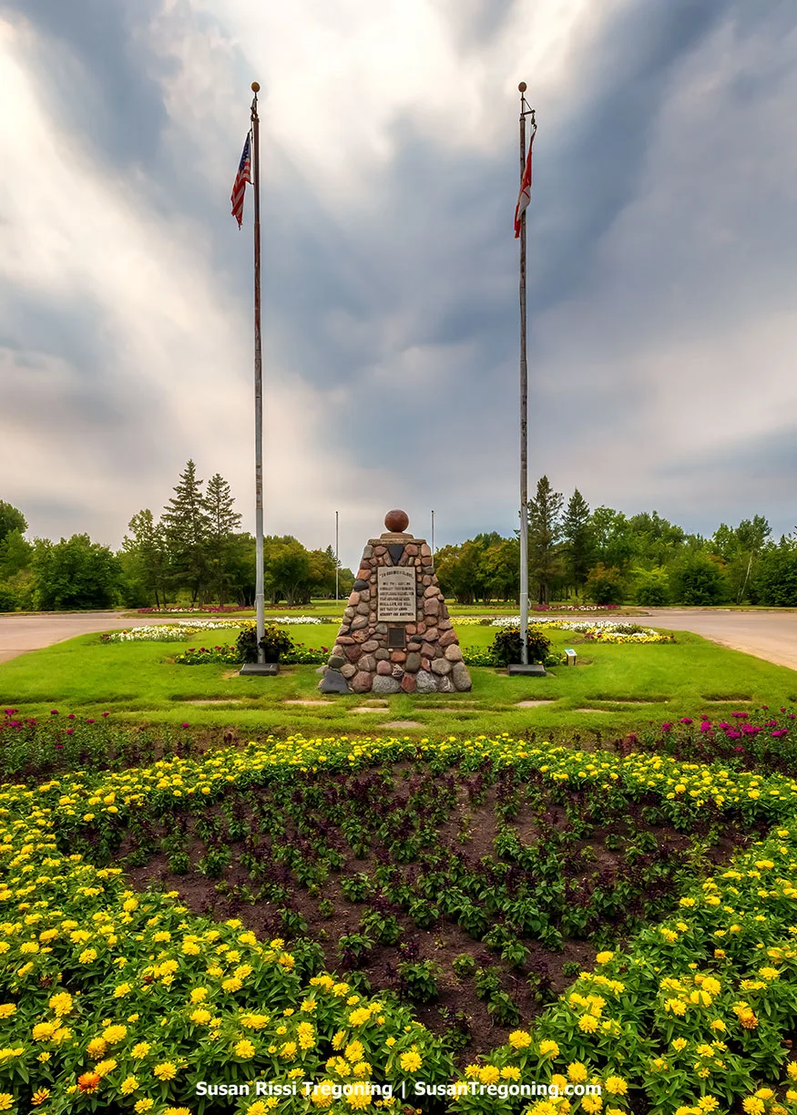 Rock Cairn Grand Opening Marker at the International Peace Garden, standing on the U.S.–Canada border between two flagpoles surrounded by vibrant yellow flowers and lush greenery under a dramatic sky.