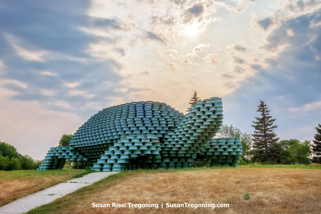 Large wheel‑rim sculpture of the W’eel Turtle near Bottineau, North Dakota, positioned atop a grassy hill along the Turtle Mountain Scenic Byway. The turtle’s rounded shell and raised head are built from stacked, turquoise‑painted steel rims, giving the sculpture a bold, textured appearance. Sitting at the crest of the hill, the landmark stands prominently against the open sky, its metal surfaces catching the light and emphasizing its playful, oversized form.